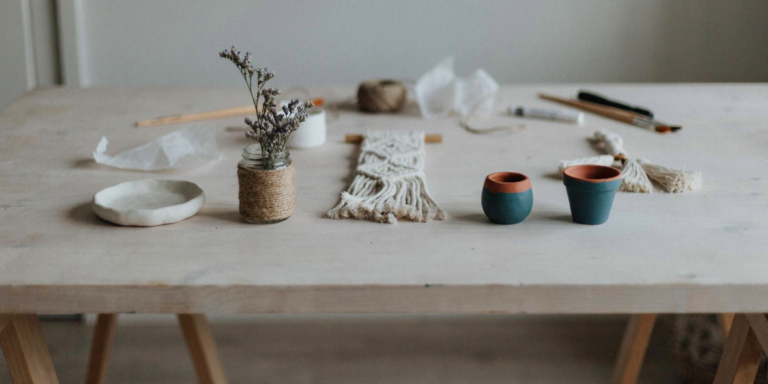 A pine worktable with several finished small crafts: a clay dish, a twine-wrapped jar, a macramé hanger and painted pots